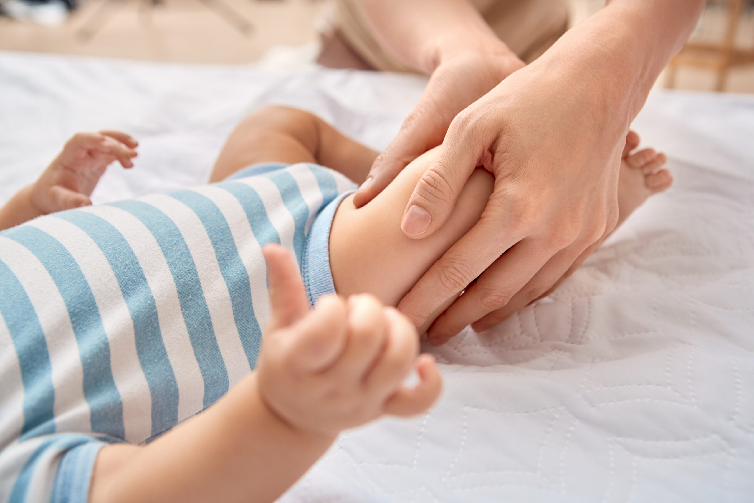Young mother taking care of little baby son wearing bodysuit lying on bed at home doing pediatric massage massaging his legs close-up